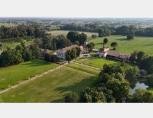 Veduta aerea di un complesso rurale con edifici in muratura e tetti in coppi, disposto attorno a cortili e viali alberati. Intorno si estendono prati e campi coltivati, con aree boscate e un piccolo specchio d’acqua ai margini.