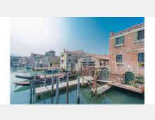 Canale con piccoli pontili in legno e pali d’ormeggio, con barche attraccate lungo la riva. Sullo sfondo si vedono edifici in mattoni e intonaco con tetti in coppi e finestre con scuri, affacciati direttamente sull’acqua.