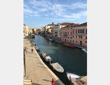 Veduta di un canale urbano con una lunga riva lastricata e piccole barche ormeggiate lungo l’acqua. Sul lato opposto si allineano edifici storici dai colori pastello con tetti in coppi e finestre ad arco sotto un cielo parzialmente nuvoloso.