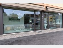 Vetrina di un negozio moderno di arredamento con grandi finestre e porta in vetro, tende a rullo e insegna verticale “INGA”. All’interno si vedono divani e tavolini esposti, con riflessi della strada e degli edifici circostanti sul vetro.