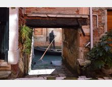 Stretto canale tra edifici in mattoni e intonaco, con un piccolo passaggio ad arco sotto una trave scura e una rampa in pietra che scende verso l’acqua. Si vedono muri usurati, tubazioni a vista e piante in vaso lungo i bordi.