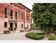 Facciata di una villa storica a due piani con intonaco rosso e infissi verdi, ingresso con pensilina in ferro e panchine su un vialetto di ghiaia. Accanto si vede un giardino con siepi basse, aiuole fiorite e grandi alberi che ombreggiano l’area.