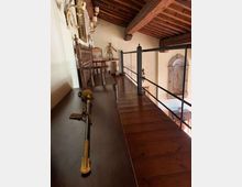 Soppalco interno con pavimento e soffitto a travi in legno, affacciato su un ambiente sottostante tramite ringhiera in metallo. Lungo la parete ci sono mobili antichi e oggetti decorativi, mentre sul muro di fronte si vede un affresco incorniciato.