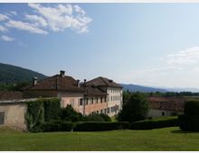 Complesso di edifici rurali con facciate chiare e tetti in coppi, circondato da prato e siepi, con alcune pareti coperte da edera. Sullo sfondo si vedono colline e montagne sotto un cielo azzurro con nuvole sparse.