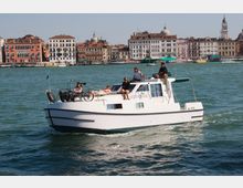 Barca a motore bianca che naviga su un ampio canale, con l’acqua increspata in primo piano. Sullo sfondo si vede un lungofiume urbano con palazzi storici dai tetti in coppi, cupole e un campanile, oltre a diverse imbarcazioni lungo la riva.