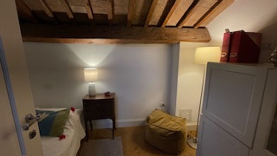 Piccola camera da letto mansardata con soffitto basso e travi in legno a vista, pavimento in legno e pareti chiare. Sono visibili un letto singolo, un comodino con lampada, una lampada da terra e un armadio accanto a un pouf.