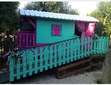 Piccola casetta in legno dai colori turchese e fucsia, con tetto ondulato bianco, finestra e veranda laterale. Davanti c’è una passerella in leggera salita con staccionata turchese decorata e fiori, circondata da alberi e vegetazione.