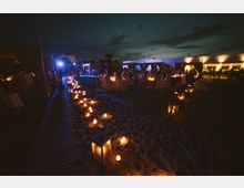 Allestimento serale su spiaggia con un sentiero di lanterne e candele sulla sabbia che conduce a tavoli apparecchiati. Sullo sfondo si vede una struttura bassa illuminata e tende bianche sotto un cielo notturno nuvoloso.