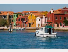 Canale con barca in primo piano e, sullo sfondo, una fila di case colorate affacciate sull’acqua con tetti in tegole rosse e finestre scure. Lungo la riva si vedono un molo e pali di ormeggio, con un lungomare davanti agli edifici.