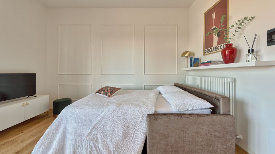 Camera da letto moderna con pareti bianche a riquadri e pavimento in legno, con un divano-letto rivestito in tessuto e biancheria chiara. A lato ci sono un mobile basso con TV e, sopra un termosifone, una mensola con libri, lampada, vaso rosso e una stampa incorniciata.