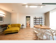 Soggiorno moderno e luminoso con pavimento in legno, divano chaise longue verde, tavolo da pranzo rotondo con sedie bianche e grande finestra con tende chiare. Sulla parete ci sono mensole in metallo e legno e una porta centrale che conduce ad altre stanze.