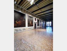 Ampia sala museale con pavimento in terrazzo e soffitto a travi, illuminata da faretti. Lungo le pareti sono esposti grandi dipinti incorniciati con stucchi decorativi, con porte laterali e alte finestre ad arco sul fondo.