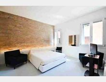 Camera da letto moderna e minimale con grande letto matrimoniale, parete in mattoni a vista e pavimento chiaro. Due ampie finestre illuminano la stanza; sono presenti una poltrona scura, una scrivania con monitor e un armadio a muro.