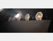 Fila di grandi fari scenici cilindrici montati su una pedana rialzata, con lenti e fori di ventilazione visibili, in un ambiente interno scuro. Un cavo e una luce intensa dall’alto illuminano parzialmente l’area tecnica.
