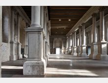 Ampia sala interna in stile classico con lunghe file di colonne in pietra e basi massicce che creano navate laterali. Soffitto a travi in legno scuro, pareti in mattoni e luce naturale che entra da finestre ad arco lungo i lati.