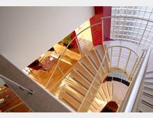 Scala a chiocciola interna con gradini chiari e corrimano in metallo, vista dall’alto su un atrio luminoso con pavimento in legno e tappeto. A lato si vede una parete curva in mattoni di vetro e un pilastro rosso che definiscono lo spazio moderno.