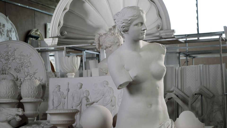 Interno di un laboratorio di scultura con grandi modelli e decorazioni in gesso, tra cui bassorilievi e una cornice a conchiglia appoggiata sullo sfondo. In primo piano si vede un busto femminile in gesso su un tavolo, circondato da altri elementi ornamentali.