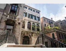 Facciate di edifici storici in pietra e intonaco affacciati su un piccolo canale, con finestre ad arco e persiane verdi. In primo piano si vede un ponte pedonale con ringhiere in ferro e gradini che scendono verso l’acqua.