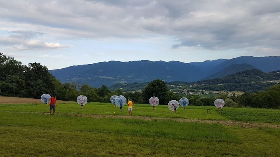 Ampio prato in collina con sullo sfondo una valle e una catena montuosa sotto un cielo nuvoloso. Nel campo sono visibili grandi sfere trasparenti distanziate lungo una linea, con alberi ai margini e terreni agricoli nelle vicinanze.