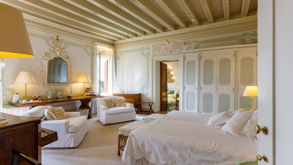 Camera da letto in stile classico con soffitto a travi dipinte e pareti decorate a motivi floreali pastello. Letto matrimoniale con biancheria chiara, armadi a pannelli, specchio ornamentale e area salotto con poltrone vicino a una grande finestra.