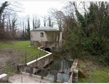 Piccolo edificio in mattoni con tetto a padiglione, parzialmente coperto di edera, accanto a un canale con una chiusa in cemento e ringhiere metalliche. L’area è immersa nel verde con prato e alberi spogli, tipica di una zona rurale o di un parco fluviale.
