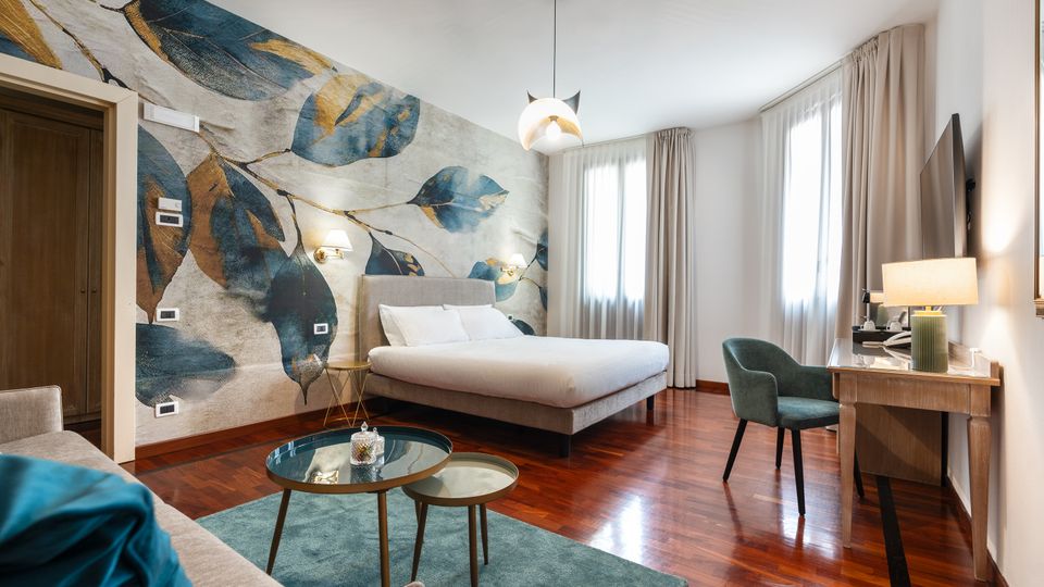 Camera da letto moderna con pavimento in legno lucido, grande letto matrimoniale e parete decorata con un murale di foglie. Due ampie finestre con tende chiare illuminano una zona scrivania con sedia e TV a parete, con tavolini bassi su tappeto verde in primo piano.