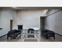 Interno minimalista con ampio soggiorno dalle pareti in intonaco grigio e pavimento lucido, arredato con due divani e una poltrona in pelle nera disposti attorno a un grande tavolino basso. Sul fondo si vedono un radiatore scuro, un mobile bianco e un corridoio laterale, illuminati da una finestra alta vicino al soffitto.