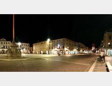 Ampia piazza cittadina di notte con pavimentazione in pietra, un alto pennone su basamento monumentale in primo piano e una lunga fila di edifici storici con portici illuminati. Sullo sfondo si vede una torre con orologio e lampioni che illuminano la strada lungo il perimetro della piazza.