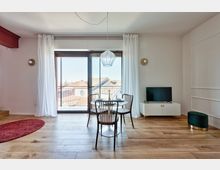 Soggiorno moderno con pavimento in legno, tavolo rotondo con quattro sedie e lampada a sospensione, affacciato su una grande porta-finestra con tende bianche che dà su un balcone con vista sui tetti. A destra ci sono un mobile basso con televisore e un piccolo pouf, con pareti bianche decorate da applique rotonde.