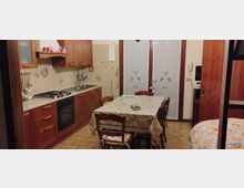 Cucina abitabile con mobili in legno, piano cottura e forno incassato lungo la parete sinistra, lavello e pensili. Al centro c’è un tavolo rettangolare con tovaglia e sedie, con pavimento in piastrelle e porte a vetro sul fondo.