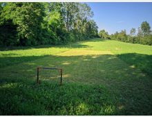 Ampio prato erboso in una radura, circondato da alberi e vegetazione fitta ai lati, con cielo sereno sullo sfondo. In primo piano si vede una piccola porta da gioco e, più lontano, alcuni paletti che delimitano un’area sul terreno.