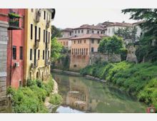 Canale urbano con acqua calma che scorre tra edifici storici dai colori pastello sulla sinistra e una riva verde con alberi sulla destra. Sullo sfondo si vedono case con tetti in coppi e un edificio con loggiato ad archi affacciato sul canale.