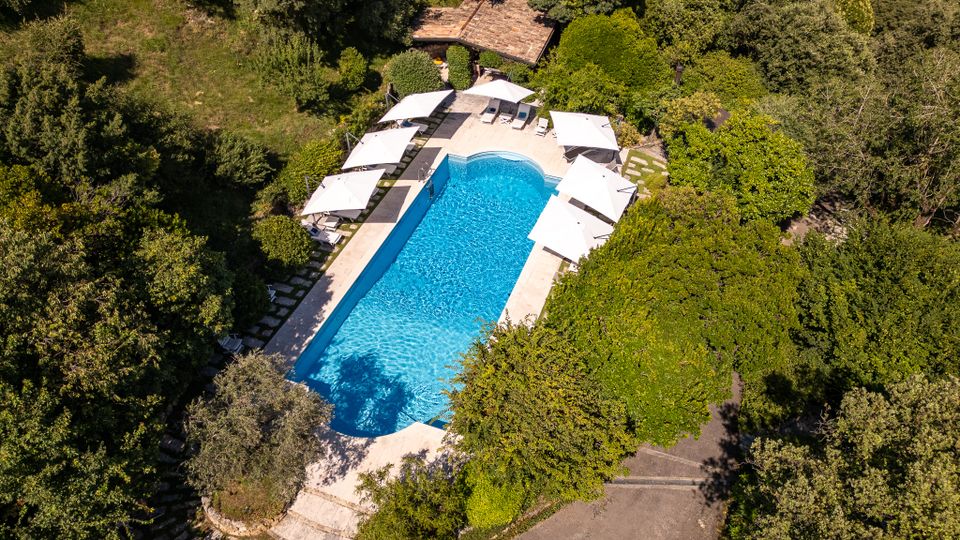 Vista aerea di una piscina all’aperto a forma rettangolare con estremità arrotondate, circondata da una terrazza con ombrelloni bianchi e lettini. L’area è immersa nel verde di alberi e siepi, con un piccolo edificio dal tetto in tegole vicino al bordo della piscina.