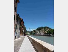 Passeggiata lungo un fiume con parapetto in pietra e lampione, affiancata da edifici storici sul lato sinistro. Sulla sponda opposta si vedono case su una collina con cipressi e vegetazione sotto un cielo sereno.