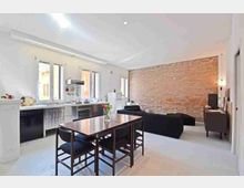 Interno open space con cucina lineare a parete sotto una fila di finestre, dotata di elettrodomestici in acciaio e piano di lavoro bianco. In primo piano c’è un tavolo da pranzo con sedie scure e, sullo sfondo, una zona soggiorno con divani neri e una parete in mattoni a vista.