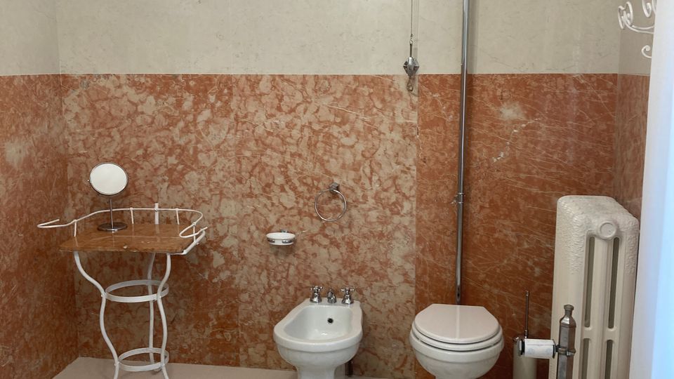 Bagno in stile vintage con pavimento a mosaico geometrico a stelle e pareti rivestite in marmo rosato e beige. Sono visibili bidet e WC con cassetta alta a tirante, un piccolo tavolino in ferro e un termosifone a colonna.