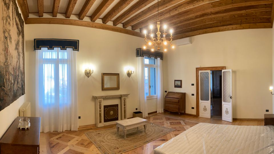 Ampia camera da letto in stile classico con soffitto a travi di legno a vista, pavimento in parquet a intarsi e lampadario centrale. La stanza include un letto con testata decorata, due finestre con tende leggere, un camino ornamentale, un arazzo a parete e una porta a doppio battente che conduce a un’altra stanza.