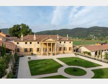 Villa storica color ocra con tetto in coppi e portico centrale a colonne, affacciata su un ampio cortile di ghiaia con aiuole geometriche di prato. Sullo sfondo si vedono campi e colline verdi, con alcuni edifici rurali ai lati del complesso.