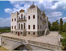 Grande villa storica bianca con basamento in pietra e tetto a cupola, caratterizzata da merlature, finestre alte e balconi con ringhiere in ferro battuto. L’edificio si affaccia su un ampio terrazzo pavimentato con balaustre, circondato da alberi e colline sotto un cielo parzialmente nuvoloso.