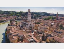 Veduta aerea di un centro storico con fitti tetti in laterizio lungo un fiume e una grande chiesa in pietra con alto campanile centrale. Sullo sfondo si vedono colline alberate e altri edifici storici con torri.