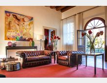 Salotto luminoso con divani Chesterfield in pelle su tappeto rosso, grande quadro colorato sulla parete bianca e soffitto con travi in legno. Ampie finestre con tende leggere e porta ad arco aperta verso l’esterno, con tavolino e vaso di fiori in primo piano.