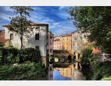 Veduta di un canale cittadino con un ponte in pietra ad archi, fiancheggiato da edifici storici dai toni chiari e tetti in coppi. Vegetazione e alberi costeggiano le sponde e l’acqua riflette le facciate sotto un cielo parzialmente nuvoloso.