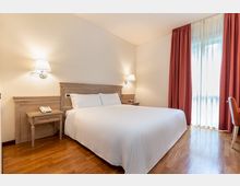Camera da letto d’albergo con letto matrimoniale, testiera e comodini in legno e applique a parete. Pavimento in parquet e grande finestra a tutta altezza con tende rosse e velo bianco che lascia entrare luce naturale.
