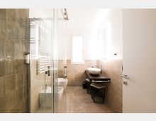 Bagno moderno e luminoso con pareti piastrellate beige e box doccia in vetro a sinistra, sanitari lungo la parete e un lavabo a ciotola su mobile in legno scuro. In fondo c’è una finestra e sul lato destro uno specchio con illuminazione.