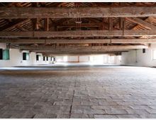 Ampio interno tipo fienile o magazzino con pavimento in pietra e pareti bianche, illuminato da finestre laterali. Il soffitto è a capriate di legno a vista con travi massicce e copertura in tegole.