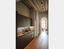 Cucina moderna e lineare in un ambiente stretto, con mobili neri opachi, lavello e piano di lavoro illuminato da luce sottopensile. Soffitto alto con travi in legno a vista, pavimento in parquet e arco che conduce a una stanza luminosa con grande finestra.