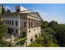 Edificio storico in stile neoclassico con facciata a portico di colonne e frontone scolpito, costruito su un basamento in pietra sopra una scarpata. Intorno si vede un giardino con prato, alberi e una statua, con bosco e cielo azzurro sullo sfondo.