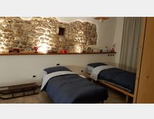 Camera da letto con due letti singoli e biancheria blu, affiancati a una parete con pietra a vista illuminata. Sopra i letti corre una mensola in legno con piccoli oggetti e decorazioni, con tenda chiara sul lato destro.