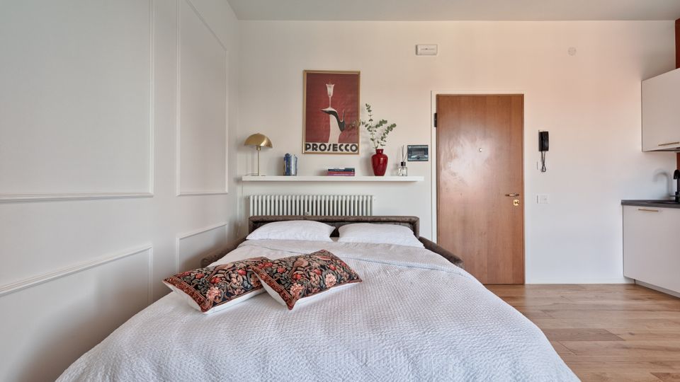 Camera da letto moderna e luminosa con letto matrimoniale, pareti bianche con boiserie e pavimento in legno. Dietro al letto ci sono una mensola con lampada e libri, un poster, un radiatore e una porta d’ingresso; a destra si vede un angolo cottura con pensili bianchi e piano di lavoro scuro.