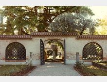 Ingresso di un giardino recintato con muro in intonaco e mattoni, copertura in coppi e portale ad arco con cancelli in ferro aperti. Ai lati ci sono due grandi aperture ad arco con griglie a losanghe, mentre all’interno si vede un vialetto di ghiaia tra alberi e una fontana centrale.